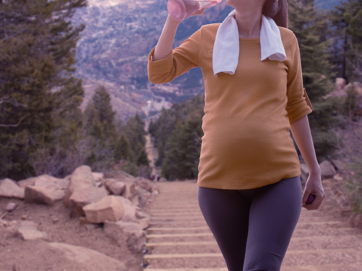 Pregnant woman hiking on a trail drinking from a water bottle.
