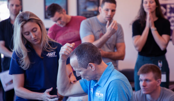 Photo of a female ART instructor demonstrating an ART protocol on the elbow of a male ART instructor as providers and students watch and practice on themselves.